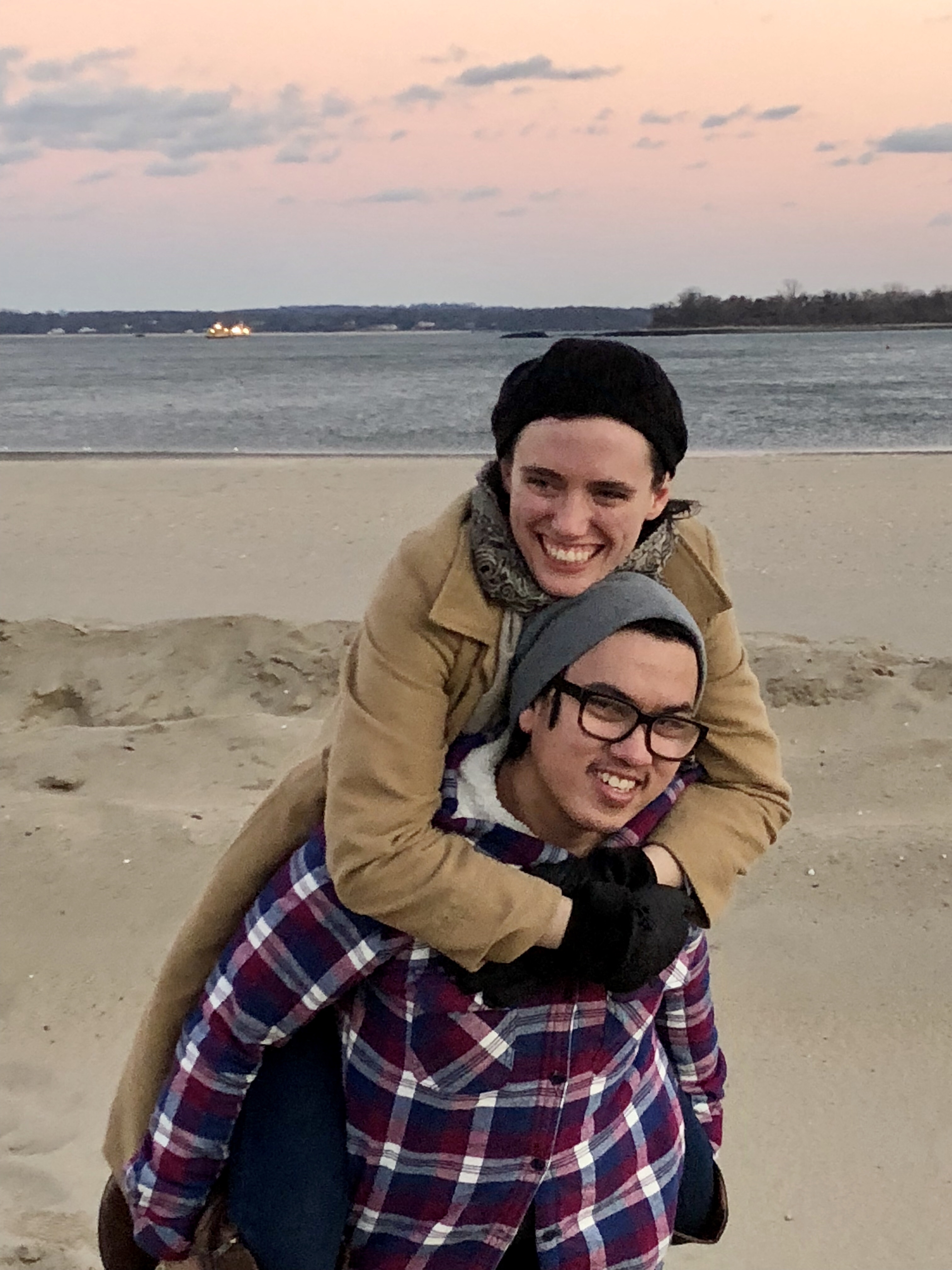 Tori (the author) clinging onto her husband’s (Johnny) back as he carries her over the sand at orchard beach in the Bronx. The sun is setting over the water, the sky is pink. They are both wearing coats and hats. Both are grinning and looking away from the camera in opposite directions. 